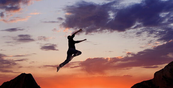 Background is sunset time. Top of picture is dark blues and purples, and ir fades to the bottom which is bright orange and reds. In the middle is a person jumping left to right, from one ledge to the other, with a seemingly large space between ledges.